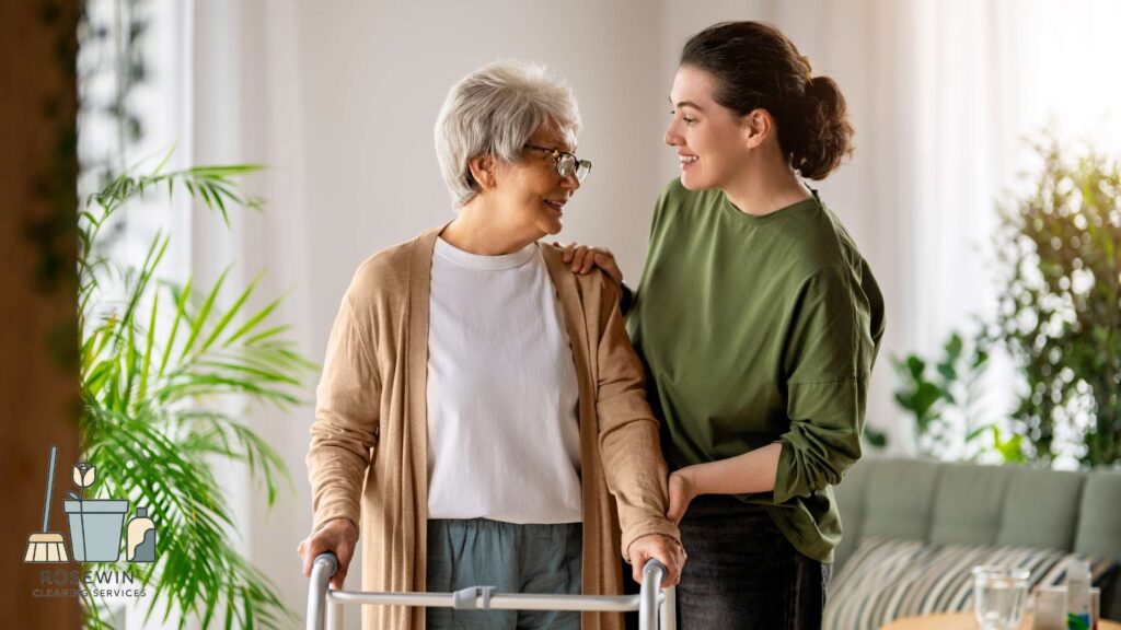 A well-lit photograph showing a smiling young woman (wearing a green shirt) supporting an elderly woman with short grey hair and glasses (wearing a tan cardigan) who is using a metal walker. They are both looking at each other warmly in a comfortable living room environment with potted plants. The 'Rosewin Cleaning Services' logo with brooms and a plant in a pot is visible in the bottom-left corner.